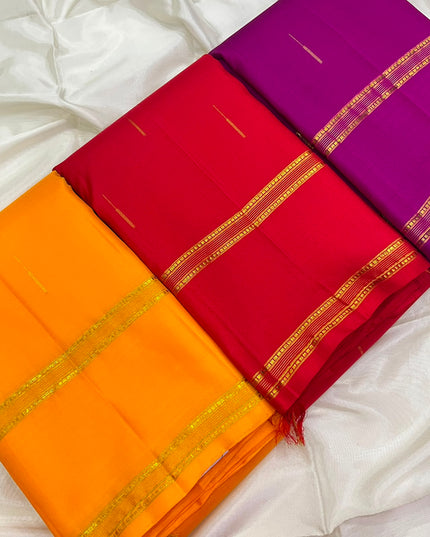 3 folded silk sarees in vibrant shades of orange, red, and purple, each decorated with a gold zari border, arranged diagonally on a white satin fabric background.
