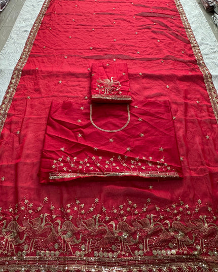 Ruby red saree laid flat with embroidered bird motifs, scattered floral details and matching unstitched blouse fabric.
